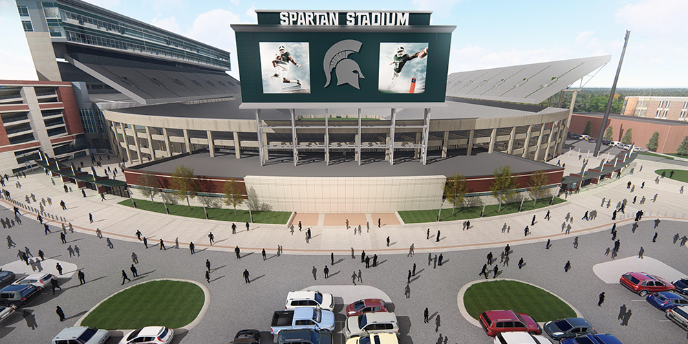 Aerial view of Spartan Stadium on a sunny day. The stadium, a testament to Granger Construction’s expertise, features a large electronic screen showcasing athletes. Enthusiastic crowds gather outside as the parking area teems with cars. Trees and walkways gracefully encircle the vibrant venue.