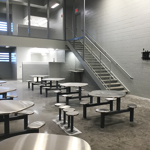 A stark, gray room with metal tables and attached benches suggests a utilitarian space, likely part of a public safety jail facility. With a staircase and railing to the right, concrete flooring, and minimal lighting, it could be in Fairfield County.