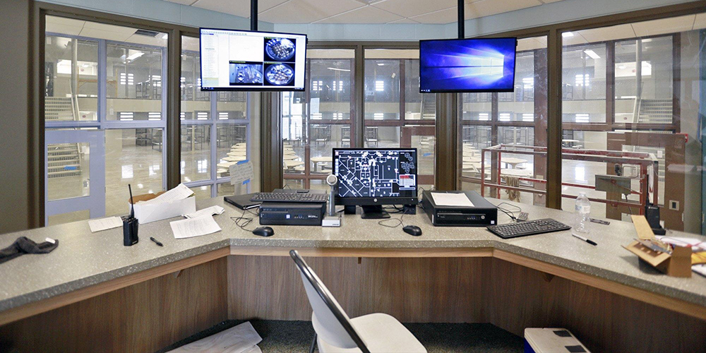 A control room in a Fairfield County public safety facility features three computer screens displaying diverse data. A swivel chair sits before the desk. Windows encircle the space, overlooking a well-lit indoor area with multiple levels and walkways.