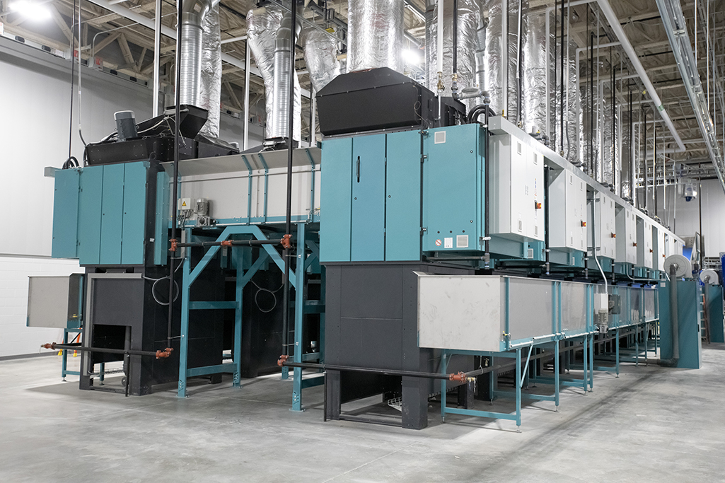 Industrial machinery with large teal and black units lined up in a spacious, well-lit facility reminiscent of an MDAHS medical laundry. Metal pipes and ductwork are visible overhead, with concrete flooring below, indicating a manufacturing or processing operation.