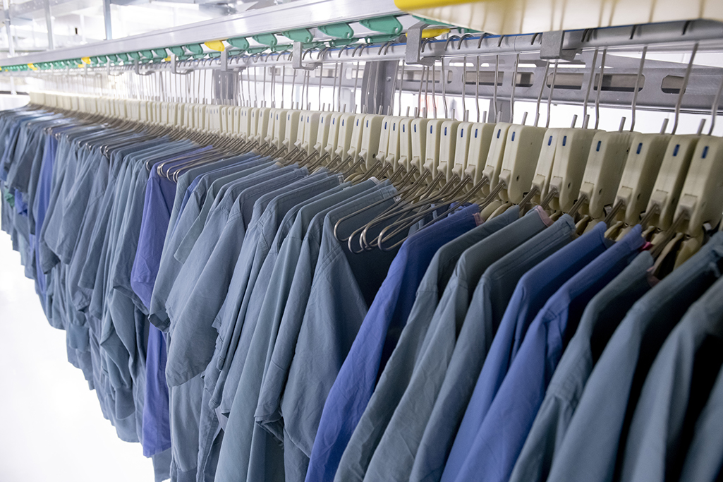 Rows of blue and green scrubs hang on metal hangers, lined up neatly on a long rail in a sterile medical laundry facility. The scrubs are meticulously organized by the MDAHS team and ready for use.
