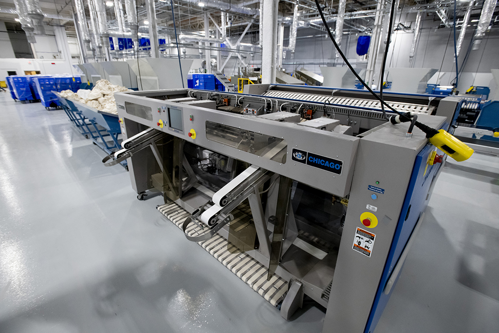 An industrial laundry machine is seen inside a large, brightly lit medical laundry facility. The machine, part of the MDAHS system, has control panels with buttons and labels. Nearby are blue carts filled with bags, and other machines are visible in the background.