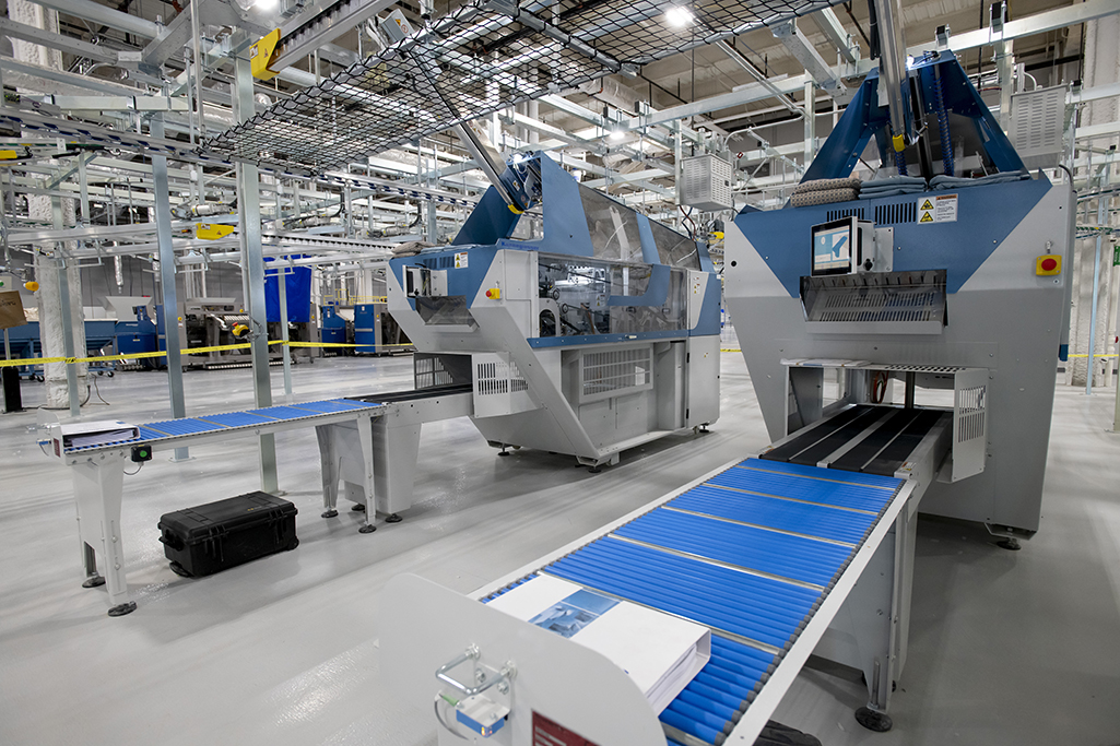 A modern industrial facility, featuring medical laundry equipment, showcases large blue and gray machinery. Two conveyor belts extend from machines under a high ceiling with pipes and cables. The area appears clean and organized.