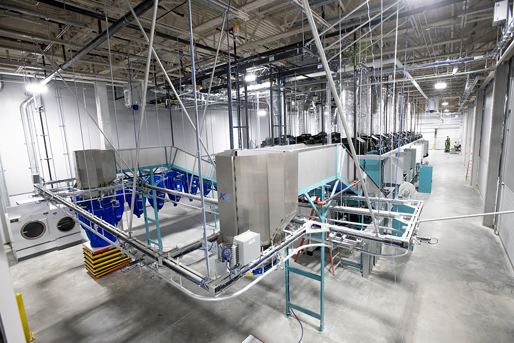 This industrial laundry facility, specializing in medical laundry, features large machinery with metal conveyors and washing units. Blue bins line the polished concrete floor beneath. High ceilings showcase exposed pipes and lights, typifying an MDAHS-compliant environment.