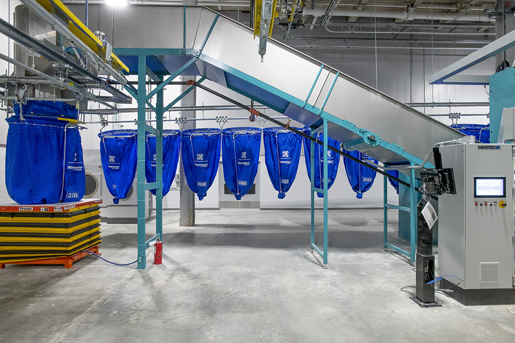 Industrial laundry facility with blue laundry bags hanging from hooks on a conveyor system. A stack of yellow pallets and a fire extinguisher are on the left, while the control panel for MDAHS operations is on the right, all on a concrete floor.