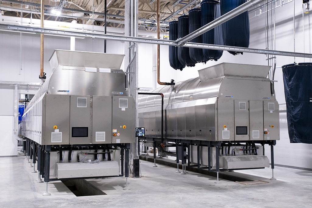 Industrial laundry equipment in a medical facility boasts two massive stainless steel washer extractor machines under a high ceiling. Ventilation ducts hang overhead, and control panels are visible on the front of these specialized medical laundry machines, ensuring hygiene and efficiency.