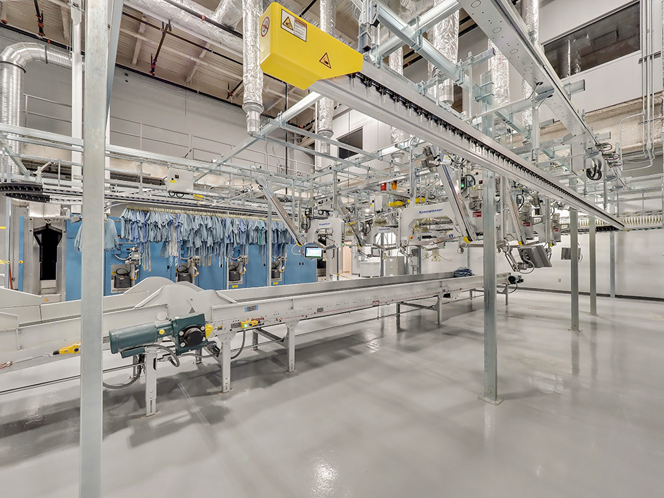 This medical laundry facility, operated by MDAHS, features automated machinery like conveyor systems and hanging racks for garments. The spacious room boasts a high ceiling, metal surfaces, and industrial lighting, creating a clean and organized environment.