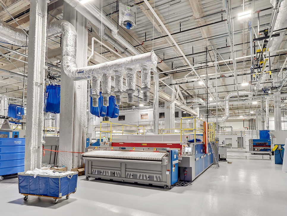 Inside the industrial laundry facility, where MDAHS services are implemented, large machinery like a folding machine dominates the scene. Blue hanging bags and metal pipes are visible among the equipment. The spotless floor contrasts with a ceiling lined with cables and fixtures overhead.