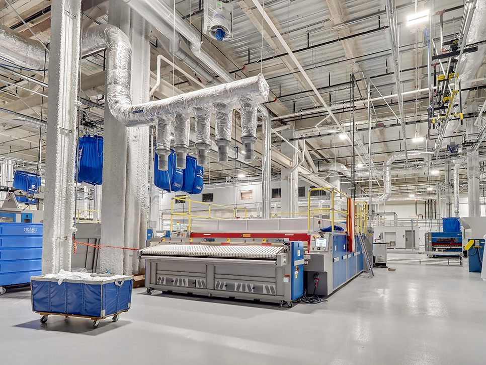 The industrial laundry facility, part of the MDAHS network, features large metal pipes and a conveyor system alongside blue machinery. Ceiling ducts are visible, with hanging blue bags for handling linens. This spacious medical laundry area is bright and efficiently organized.