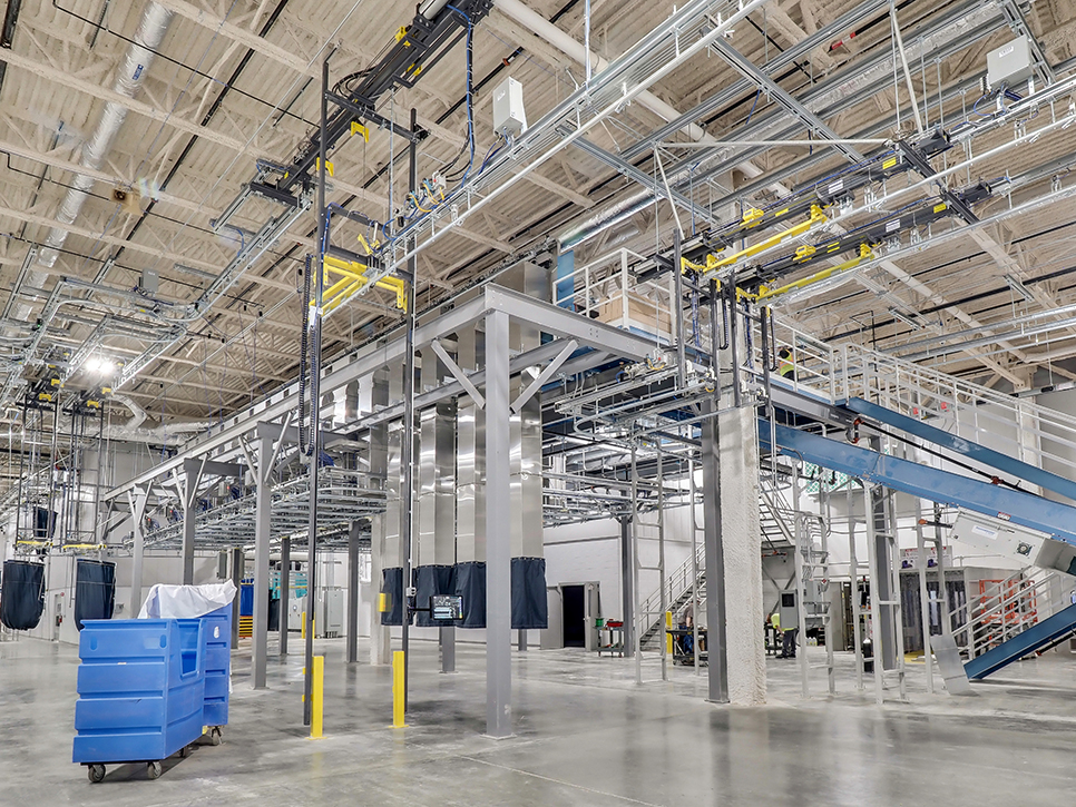 The interior of the MDAHS industrial facility features metal structures and machinery, including overhead conveyor systems. Blue bins dot the floor, while staircases lead to elevated platforms. This medical laundry space is brightly lit with overhead lights.