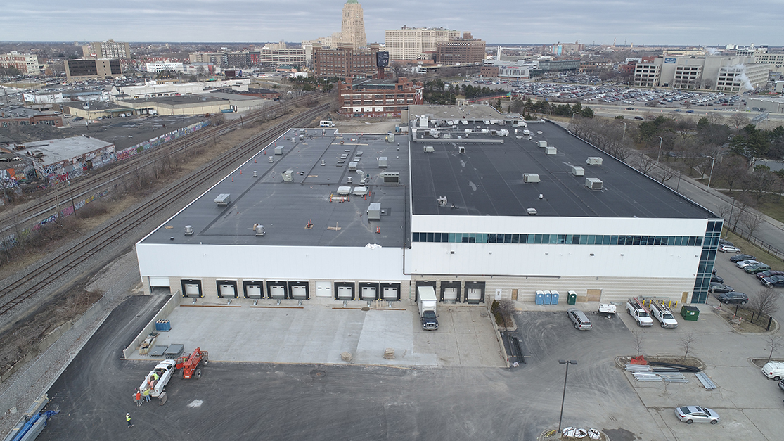 An aerial view reveals a large warehouse, potentially an MDAHS Medical Laundry Facility, with loading docks and parked vehicles. Train tracks encircle the site, set against an urban cityscape dotted with buildings beneath a cloudy sky.