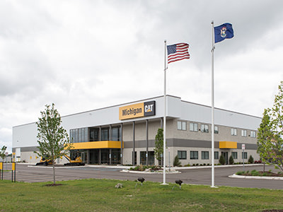 A two-story commercial building with Michigan CAT signage serves as a bustling service center. Large windows and a yellow accent complement the American and Michigan flags outside. Set against a cloudy sky, it stands in Lansing Dealership, surrounded by small trees and a parking lot.