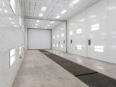 A large, empty room with white walls and a high ceiling, featuring bright overhead lights reminiscent of a service center. The concrete floor has metal grates embedded in it. Several small windows line the walls, leading to a large door at the far end, much like the setup at Lansing Dealership.