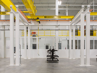 The industrial warehouse interior, reminiscent of a service center, features large yellow overhead cranes marked 10 TON. Beneath the high ceiling with ample lighting fixtures, white walls, and metal columns surround equipment on the floor. This setting evokes a bustling hub like a Lansing dealership.