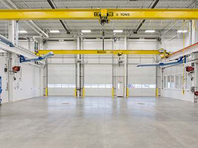 Inside the spacious interior of an empty industrial warehouse, which doubles as a Michigan CAT service center, high ceilings tower above. Yellow overhead cranes marked 3 tons span the space, while large garage doors and bright lighting illuminate the concrete floor. Blue and red machinery lines the walls.