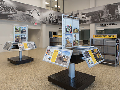 An indoor showroom at the Lansing Dealership displays heavy machinery information panels, showcasing images and details of construction equipment. The space features large wall graphics of excavators, with areas labeled Sales • Rental and Genuine CAT® Parts, all part of Michigan CATs Service Center experience.