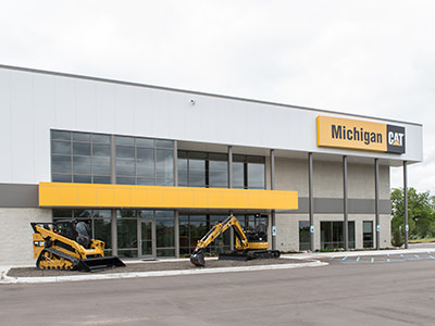 The exterior of the Lansing Dealership for Michigan CAT features large windows and a prominent yellow sign. In front, two heavy machinery vehicles stand proudly under an overcast sky, hinting at the reliable service center inside.