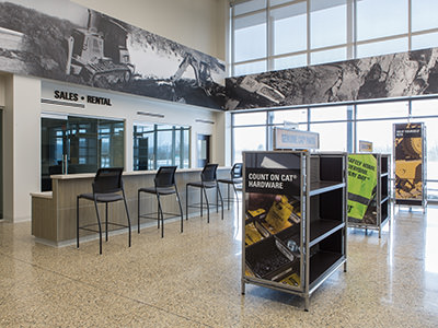 The interior of the Lansing Dealership features high stools at a counter. Large windows flood the space with natural light, while displays reading Count on Cat Hardware and safety signs catch your eye. A black-and-white mural of construction equipment adorns the wall, highlighting its role as a premier Michigan CAT Service Center.