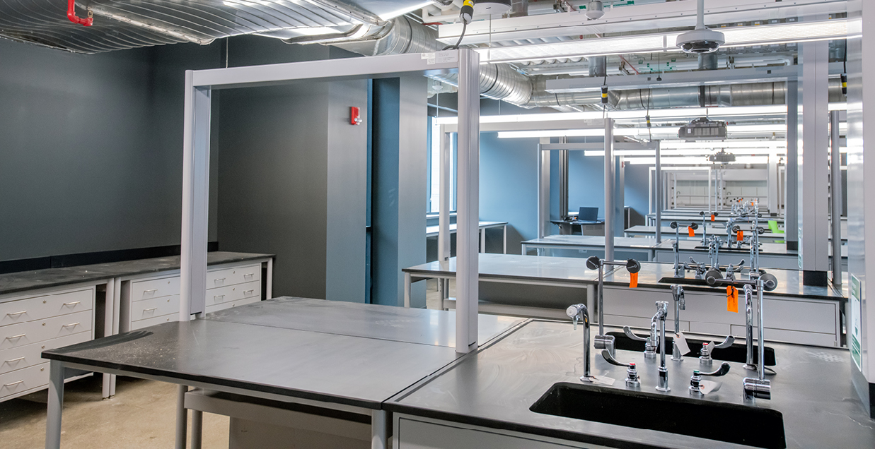 A modern laboratory at Wayne State Universitys Innovation Learning Center, featuring stainless steel tables, faucets, and drawers. The space showcases empty workstations and ventilation ducts on the ceiling in a gray and blue color scheme, embodying cutting-edge STEM design.