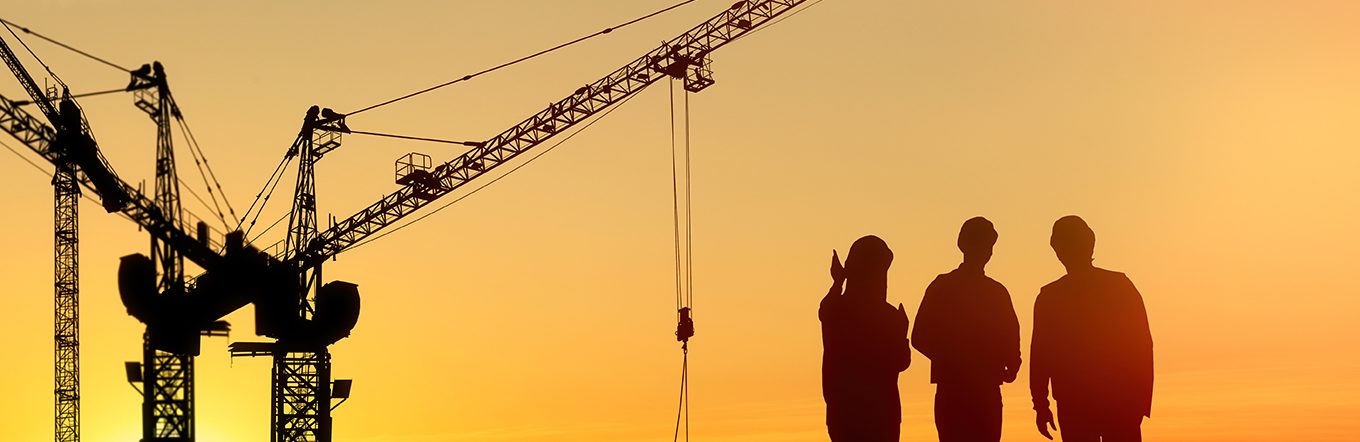 Silhouetted against a sunset, three people wearing construction helmets stand together, observing several cranes at a construction site. The Ohio Team gazes forward, the sky a gradient of warm orange and yellow hues, as new leadership steers them toward growth.