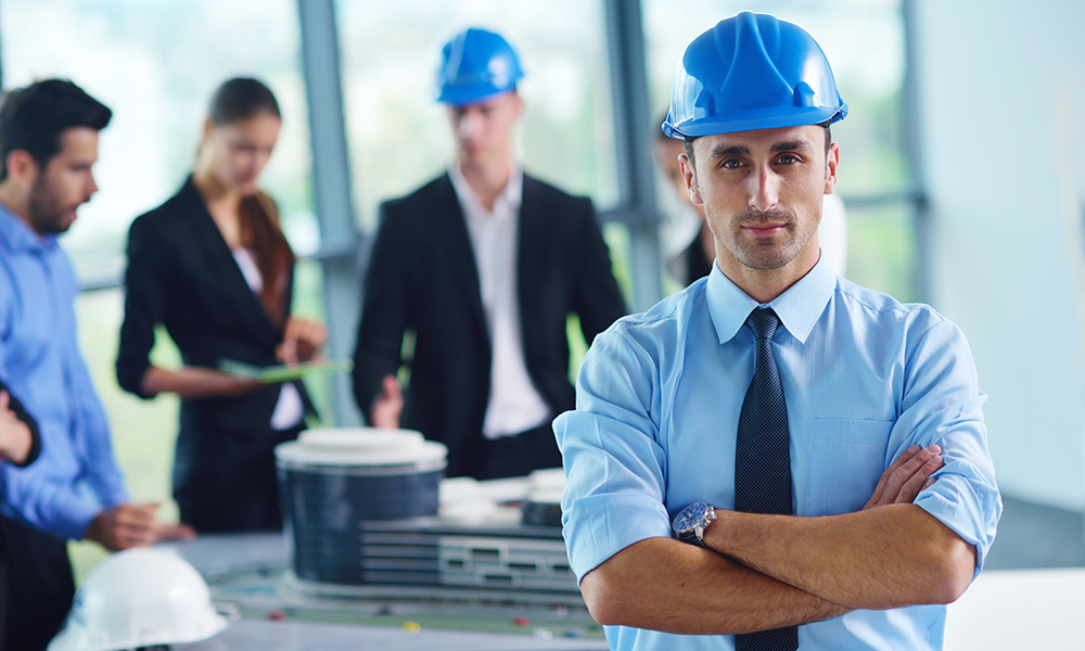 A confident man in a blue shirt and hard hat stands with arms crossed in the foreground, embodying career advancement. In the background, a group of people in business attire discuss plans near a table with blueprints, showcasing the dynamic world of construction careers in a bright, modern office.