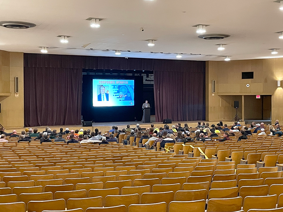 A person stands at a podium on stage, presenting to a sparsely filled auditorium. A large screen displays slides about corporate reports and company values. Rows of empty wooden seats dominate the foreground, highlighting the need for more involvement.