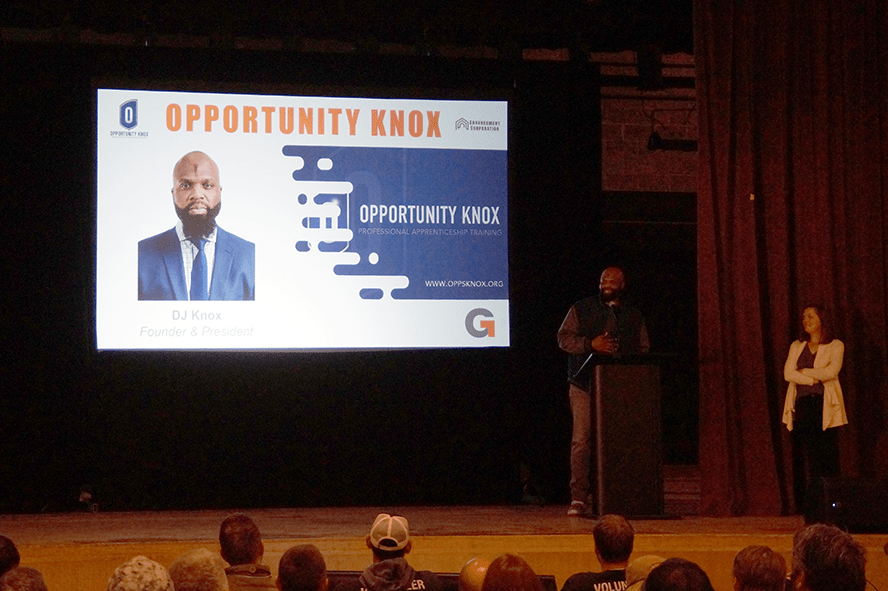 A man stands at a podium addressing an audience in a dimly lit room. Behind him, a large screen displays Opportunity Knox alongside the image of a man in a suit. Emphasizing company values, a woman stands beside the podium, attentively observing his message.