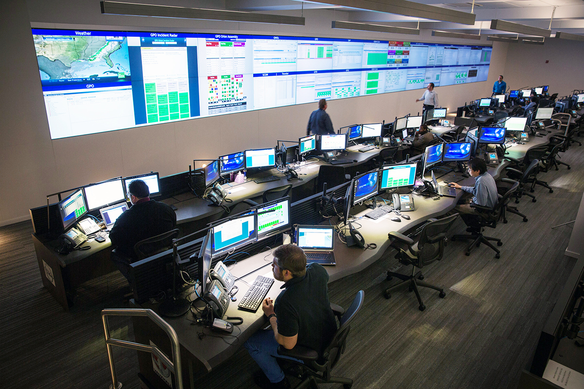 The modern control room, a testament to Granger Constructions expertise in advancing the art of building, features multiple workstations and large display screens spanning one wall. Several people are seated at desks with multiple monitors, while others stand nearby, analyzing data on screens.