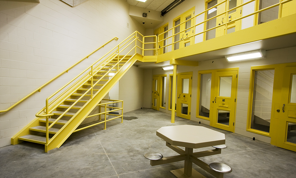 The image depicts the interior of a prison, highlighted by yellow doors and a staircase. A gray table with attached stools rests on the concrete floor under bright lights. This facility, featured in Correctional News, exemplifies innovative design by Granger using alternative project delivery methods.