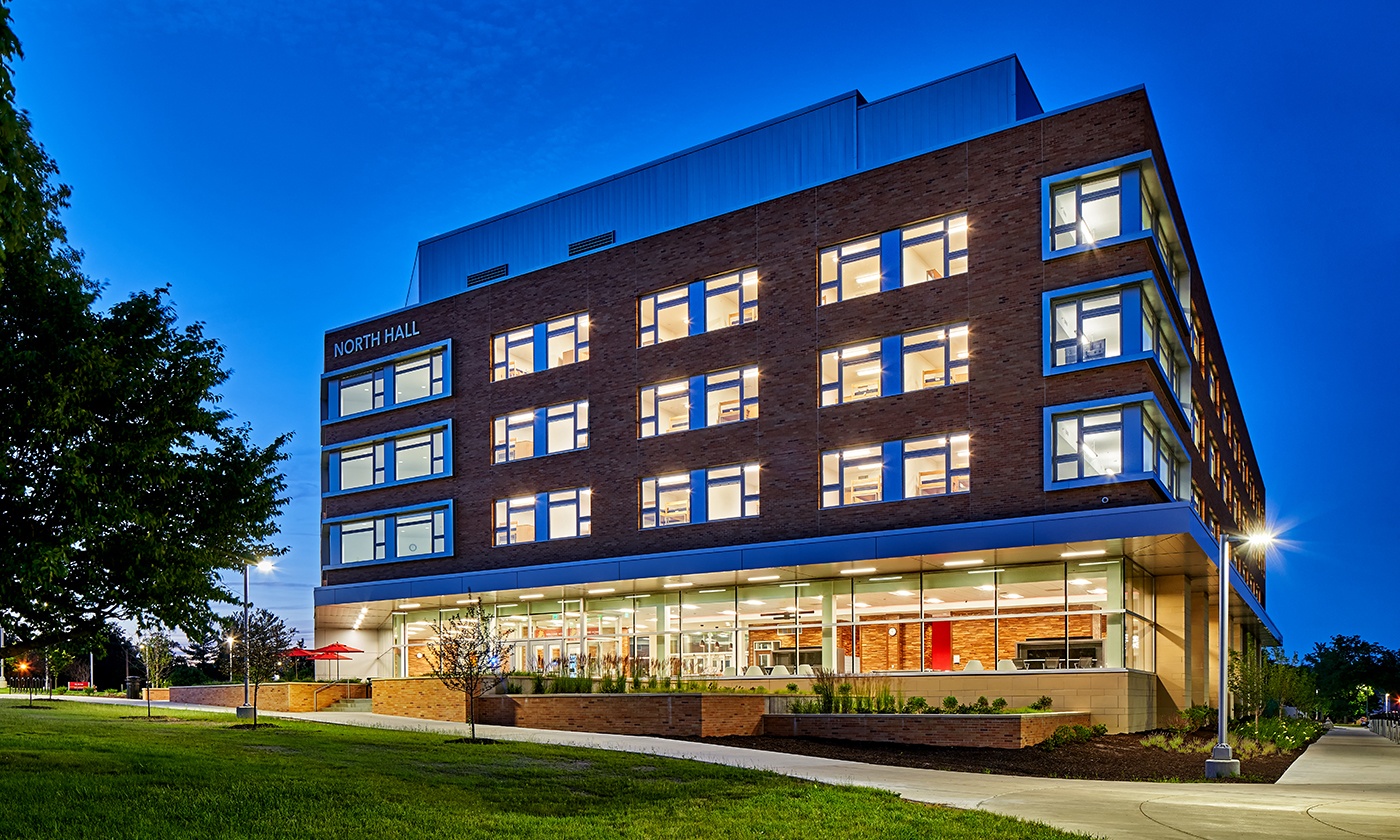 A modern, multi-story brick building labeled North Hall at Ferris State University is beautifully illuminated at dusk. Large windows reveal lit interiors. A pathway surrounds the building, with trees and grass adding to the serene scene, perfect for a campus photo gallery.