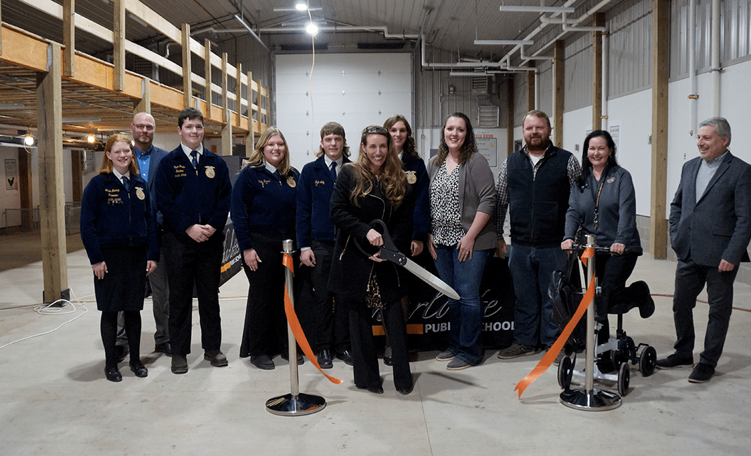 A group, including students in blue jackets and adults in various attire, gathers at the new Agricultural Learning Center. In the center, a woman with giant scissors cuts an orange ribbon. This event is a proud moment for Charlotte Public Schools, highlighted by orange ribbons and black barriers.