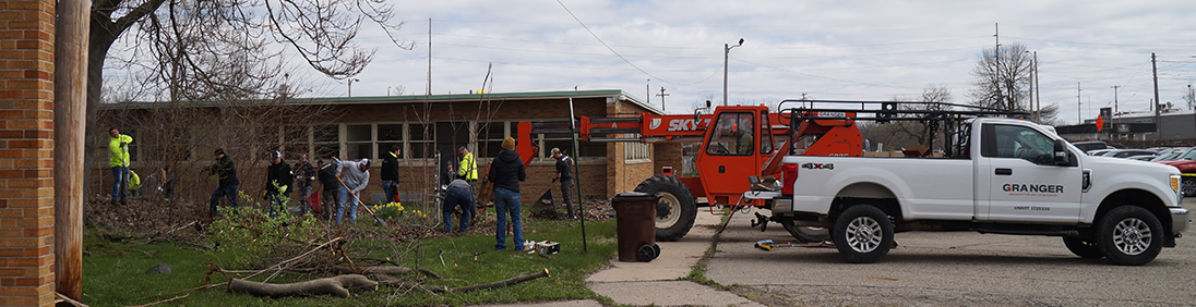 People are working outdoors near a brown building, clearing debris and branches as part of a Corporate Day of Service. An orange lift vehicle and a white pickup truck with Granger on the side are parked nearby under a cloudy sky, embodying company values in action.