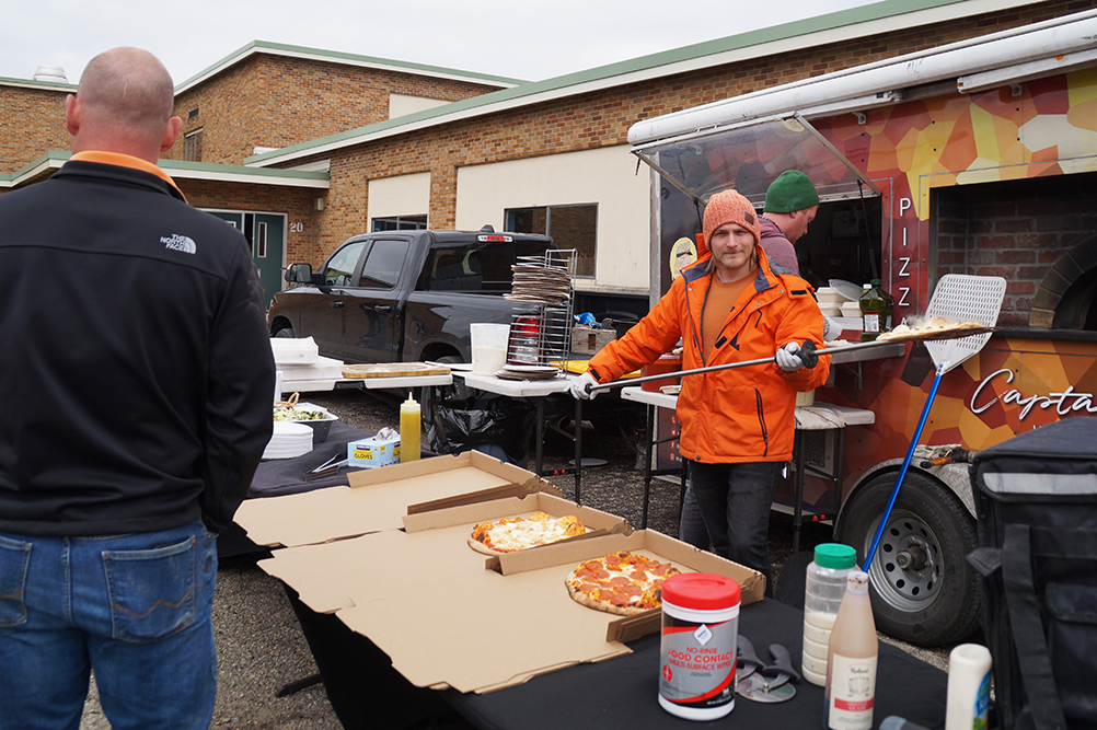A person in an orange jacket and beanie serves pizza from a colorful food truck, embodying company values of community and service. They stand by two pizzas on a table as a man in a black jacket looks on, against the backdrop of a brick building.