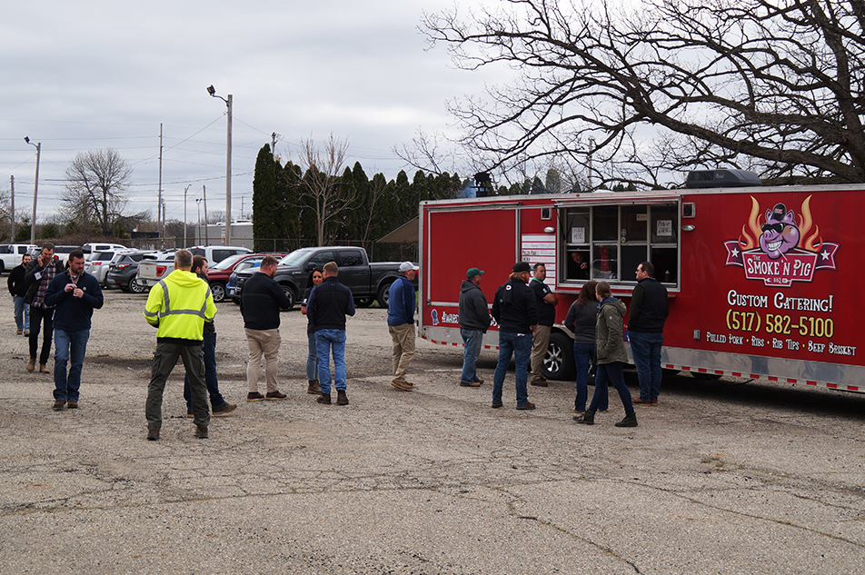 People standing in line at a red food truck during a Corporate Day of Service in a parking lot. Some are wearing jackets and hats. A few vehicles are parked in the background, and trees without leaves are visible under a cloudy sky.
