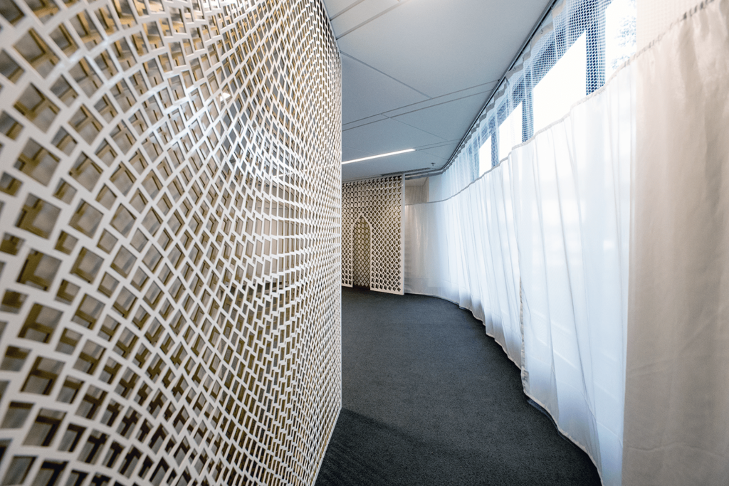 A modern hallway resembling a serene worship space, with a curved design featuring a lattice-patterned wall on the left and sheer white curtains on the right. The ceiling is fitted with lighting, and the dark gray carpeted floor compliments the natural light filtering through.