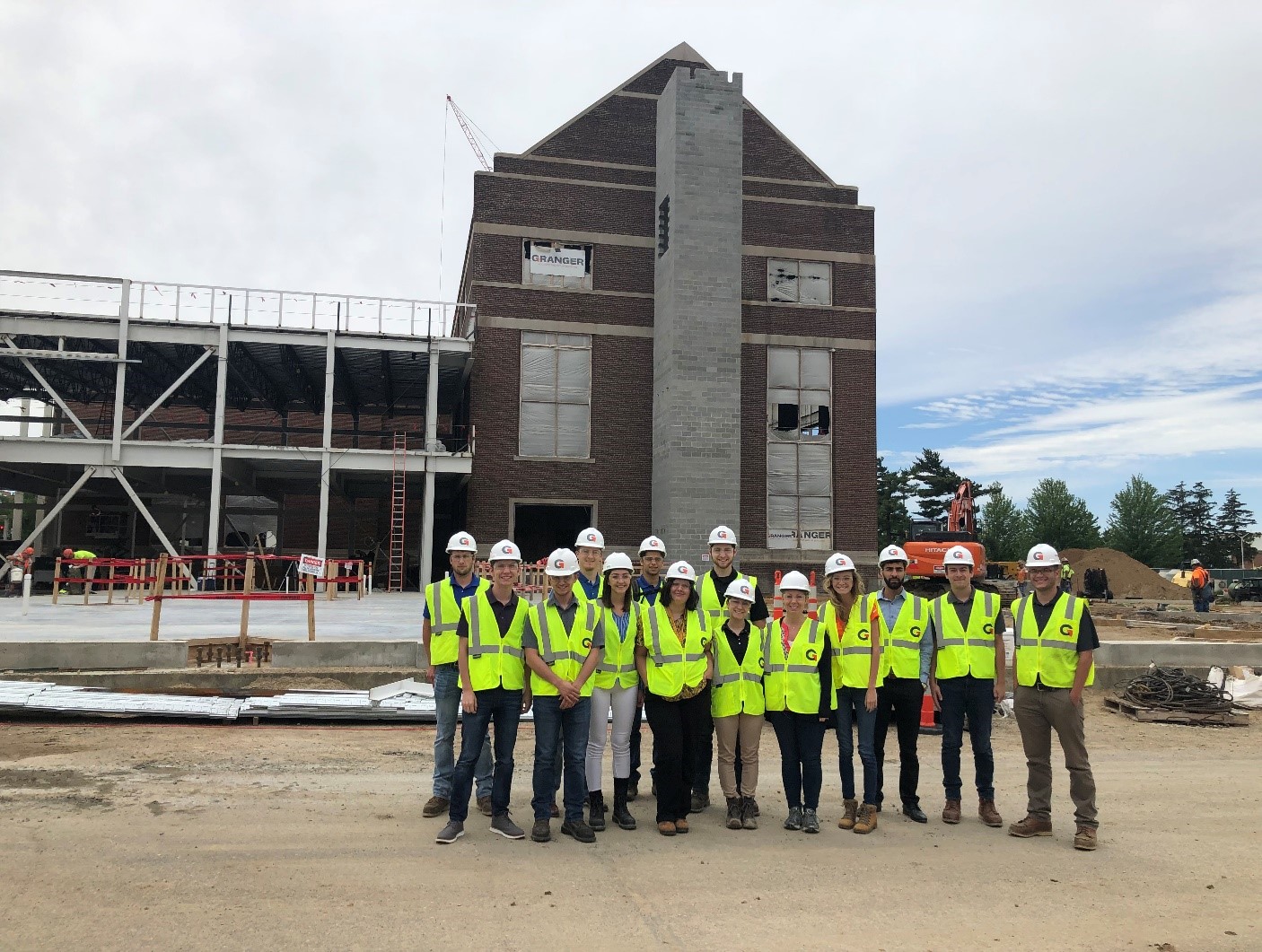 A group of people wearing hard hats and yellow safety vests stand triumphantly in front of a construction site with a partially built brick building, embodying the success of their internship program. The sky is overcast, and expanding construction equipment is visible in the background.