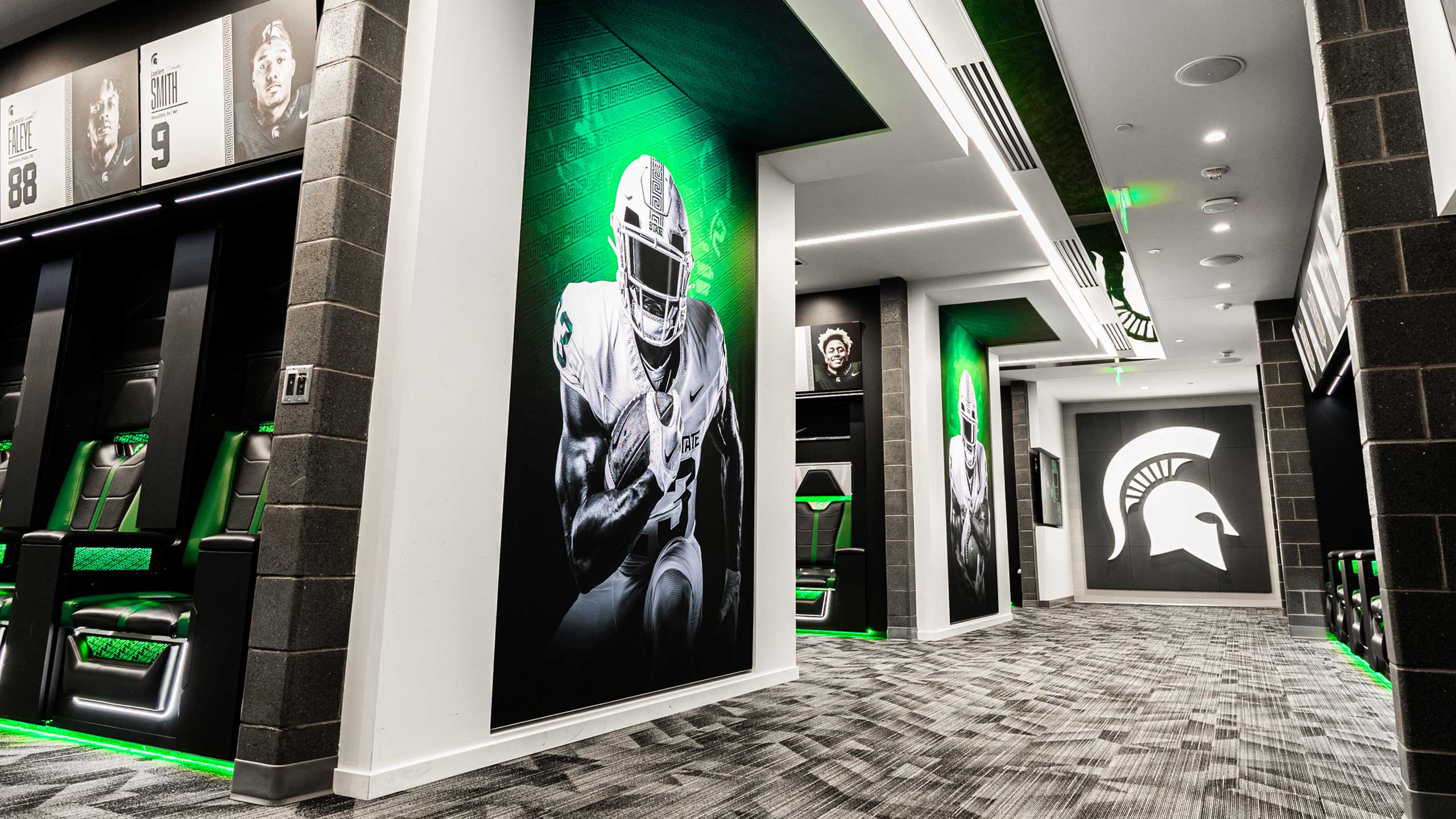 A modern sports locker room with black and green accents embodies a civic pride. The walls feature large images of football players in action, and a prominent Spartan helmet logo is displayed on one wall. Sleek, illuminated lockers complement the municipal-themed patterned carpet.