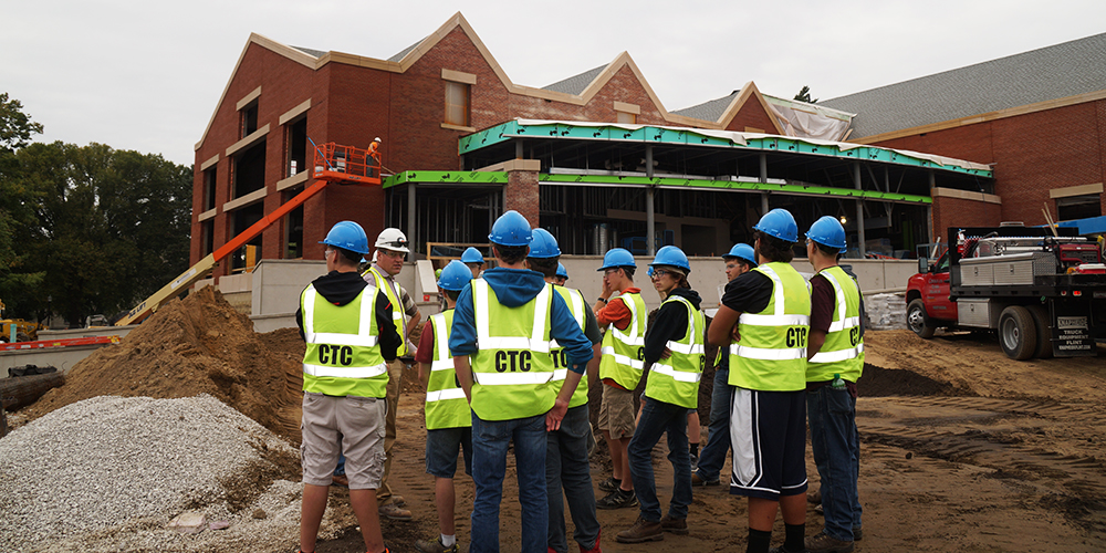 A group of people wearing blue hard hats and neon yellow vests labeled CTC enthusiastically stand at their construction job site in front of a large brick building under construction. An orange lift operates nearby, alongside piles of dirt and gravel, as new grads eagerly dive into hands-on experience.