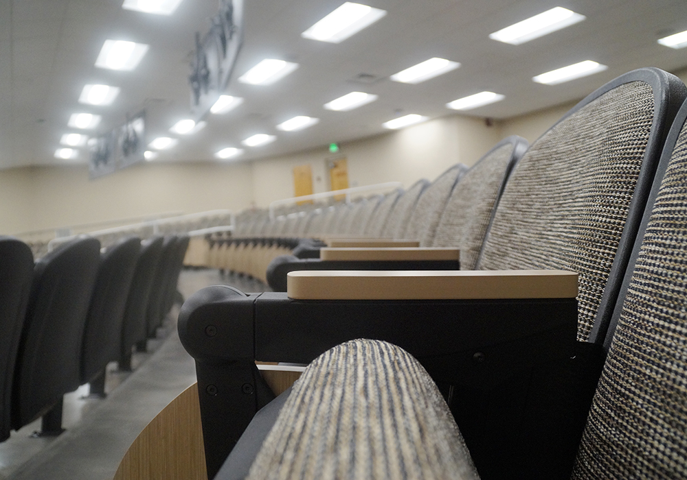 Rows of empty, beige fabric-covered seats with wooden armrests are arranged in a modern lecture hall at MSU Bessey Hall. The brightly lit room features several ceiling light fixtures, and a small set of doors is visible at the back, reminiscent of a scene from an elegant photo gallery.