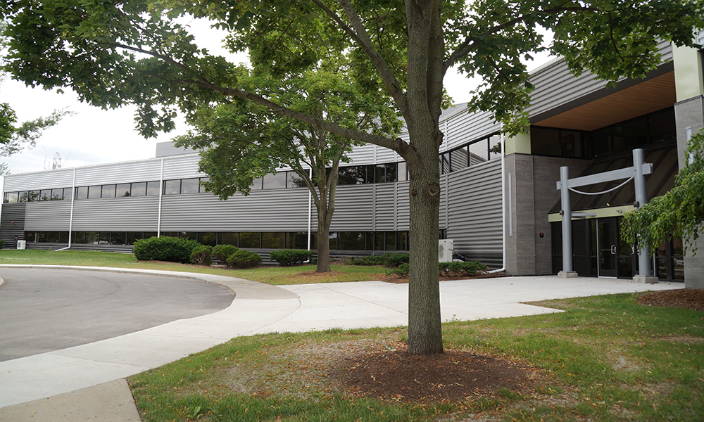 A modern office building with sleek, horizontal metal panels stands proudly in the MSU Foundation area, surrounded by trees and greenery. A curved driveway leads to the glass-doored entrance with a minimalist awning. The overcast sky sets a contemplative mood for this architectural photo gallery.