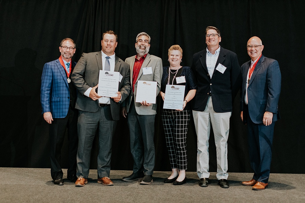 Six people stand on stage in formal attire, holding certificates. Three proudly wear medals. Their smiles light up the scene as they face the camera against a black curtain backdrop, celebrating their achievements at the MSU STEM Design Award ceremony.