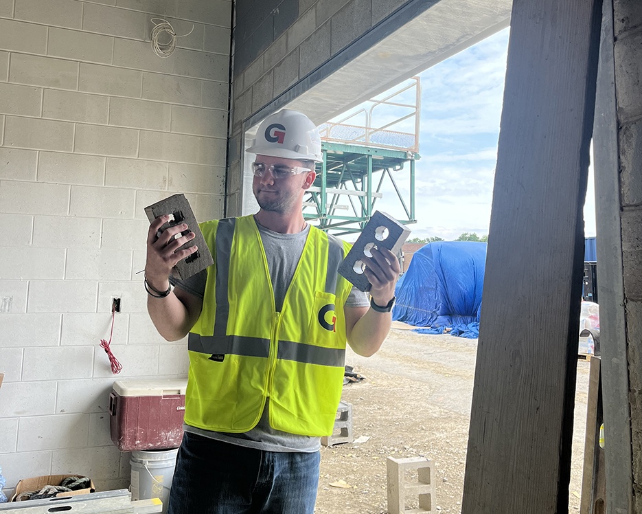 A person wearing a white hard hat and yellow reflective vest stands indoors at a construction site, holding two bricks. Alex Carroll, a Project Engineer Intern, is surrounded by the backdrop of a large blue tarp and various outdoor construction materials.