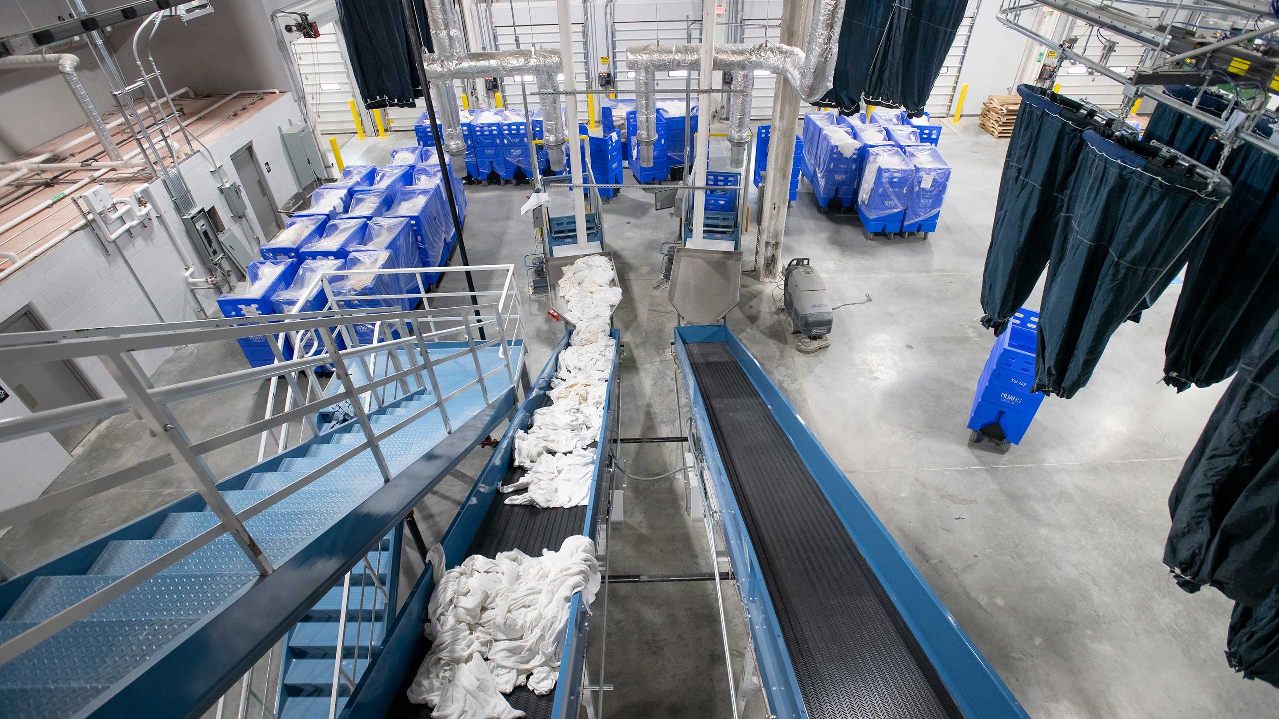 The municipal industrial laundry facility hums with efficiency. Blue bins and black conveyors transport white linens seamlessly, while empty blue carts stand ready. Overhead pipes, hanging black bags, gray flooring, and stairs complete the civic-driven operations orderly landscape.