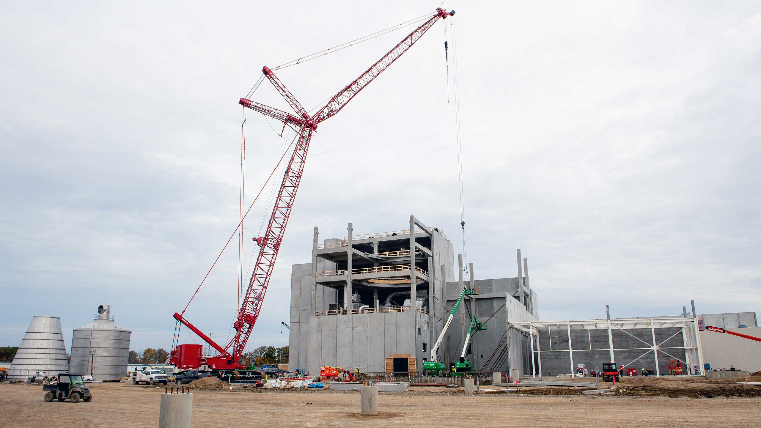 A civic construction site features a large red crane beside a towering, gray, partially-built concrete structure. Numerous small vehicles and construction equipment are visible below the cloudy sky.