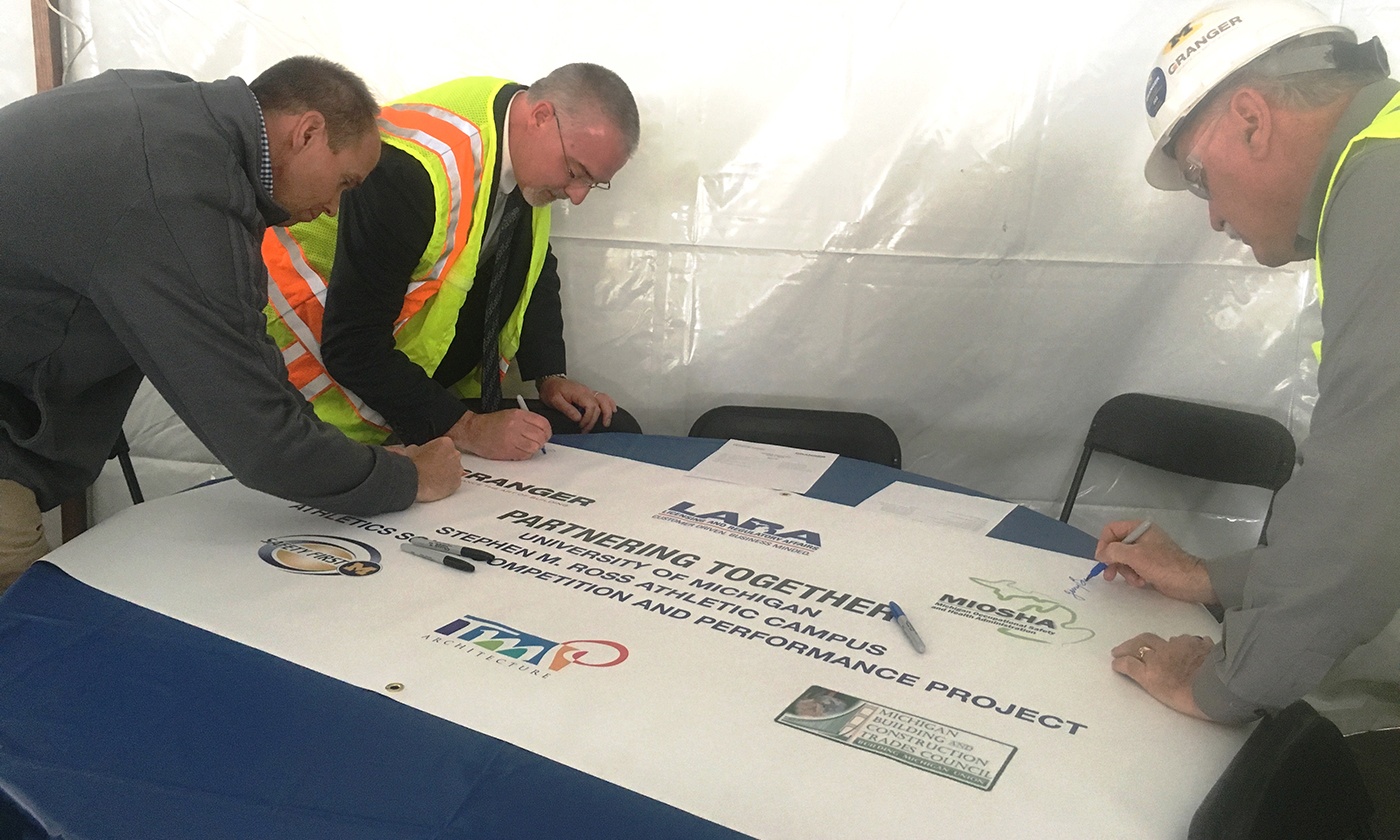 Three men in safety gear and suits are signing a large banner at a construction site. Emphasizing safety culture, the banner, adorned with various logos, reads Partnering Together, highlighting their commitment to a university athletic campus project.
