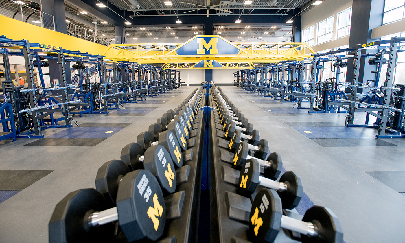 A gym with rows of dumbbells and the iconic yellow M logo from Granger Projects. Blue and yellow workout equipment lines both sides, while a large M stands prominently on a background structure. This bright, open space was recognized at the Pyramid Awards in 2017 for its design.