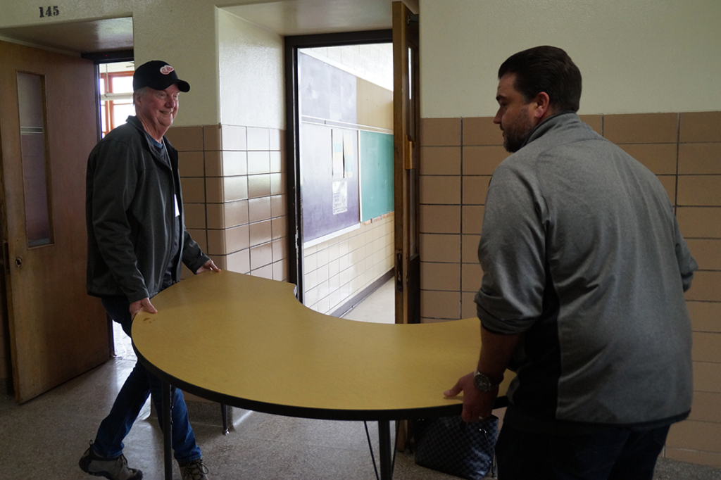 Two men are efficiently carrying a curved table through a hallway, embodying lean philosophies. The walls are tiled, with numbered room doors visible. One man wears a cap, and both are dressed casually, effortlessly reflecting company values in their teamwork.