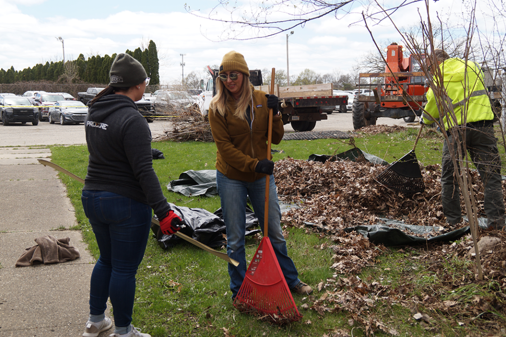 Two people are raking leaves on a grassy area beside a parking lot during a Corporate Day of Service. One wears a beanie and jacket, holding a red rake, while the other is in a cap and jacket. In the background, someone in a yellow jacket employs machinery, embodying company values.